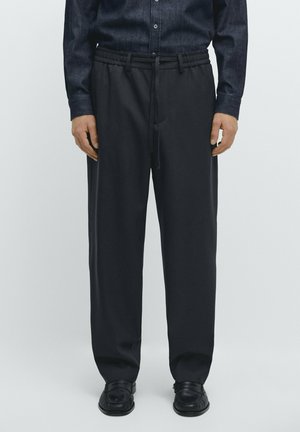 Homme portant un pantalon noir à jambes droites avec taille à cordon, des chaussures habillées noires et une chemise foncée à boutons, debout devant un fond uni.