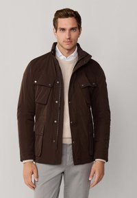 Mand iført en mørkebrun jakke med knap-lommer, beige sweater, hvid skjorte og lys grå bukser foran en ensfarvet baggrund.