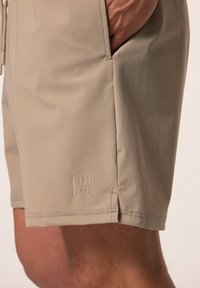 Beige zwembroek met een gladde textuur, zijzakken en een klein logo op de onderrand. Eenvoudig ontwerp met een losse pasvorm.