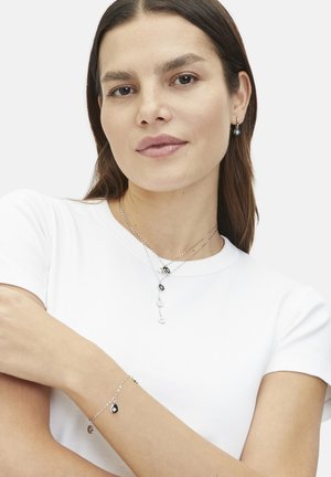 Silberne Schmuckstücke mit geschichteten Ketten, einer Mischung aus schwarzen und weißen Anhängern sowie passenden Ohrringen, die mit einem schlichten weißen Hemd getragen werden.