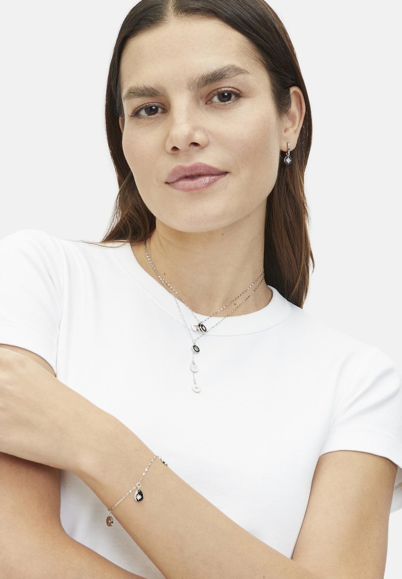 Silver jewelry featuring layered chains, a mix of black and white pendants, and matching earrings worn with a plain white shirt.