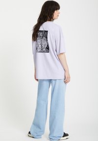 Lichtpaars oversized t-shirt met zwarte grafische print van handen op de rug; gecombineerd met lichtblauwe wijdvallende spijkerbroek en zwarte sneakers.