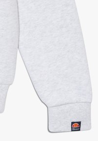 Grau sweatshirt ärmel mit geripptem bündchen, das ein kleines schwarzes etikett mit dem orange und roten Ellesse-logo trägt.