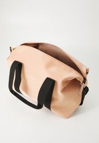Peach-farget vanntett duffelbag med svarte tekstilhåndtak og en åpning på toppen; har en subtil logo og en glatt, blank overflate.
