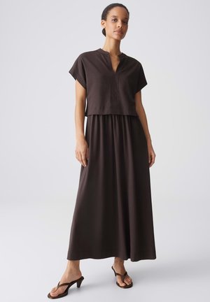 Frau steht in einem dunkelbraunen, kurzärmeligen V-Ausschnitt-Oberteil und passendem langen Rock, trägt braune Sandalen mit Absatz vor einem schlichten hellen Hintergrund.