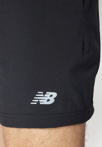 Calções atléticos pretos feitos de um tecido liso e leve. Apresenta um logo da New Balance em branco perto da barra e bolsos laterais.