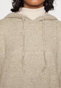 Femme portant un sweat à capuche en maille beige avec cordons sur un pull à col roulé côtelé crème, cadré sous le nez.