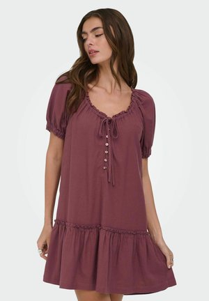 Kvinne iført en korteermet fiolett kjole med rysjekant, knapper foran og knyting ved halsen, stående foran en ensfarget bakgrunn.