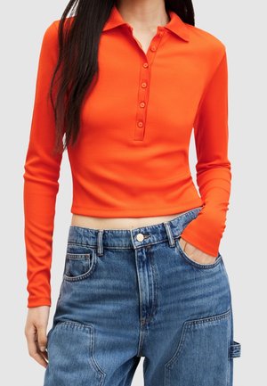 Pomarańczowa polo z długim rękawem o prążkowanej fakturze, z zapięciem na guziki, noszona z jasnoniebieskimi dżinsami z wysokim stanem, posiadającymi kieszenie z przodu.