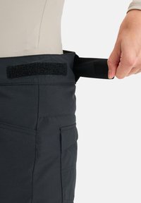 Zwarte stoffen broek met een klittenbandsluiting; hand die de verstelbare band vasthoudt. Heeft een zijzak en een gladde textuur.