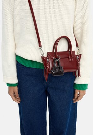 Osoba ubrana w kremowy sweter z zielonymi mankietami i niebieskie dżinsy, niosąca małą czerwoną skórzaną torebkę z przymocowaną czarną mini torebką.