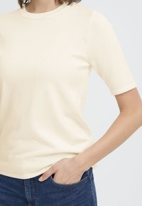 Personne portant un haut en tricot crème à manches courtes et un jean bleu, une main dans la poche avant, sur un fond clair uni.