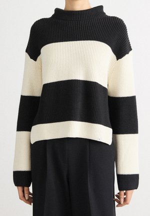 Person iført en sort og hvid bred, vandret stribet strikket sweater med lange ærmer og sorte bukser.