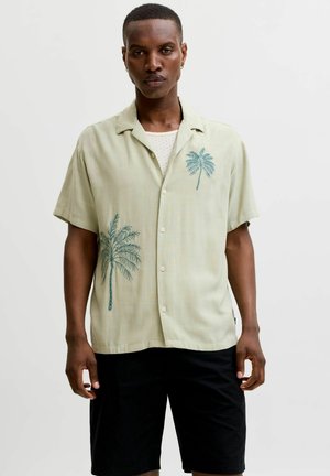 Homme portant une chemise beige clair à manches courtes avec des motifs de palmiers bleus et un short noir sur un fond clair uni.