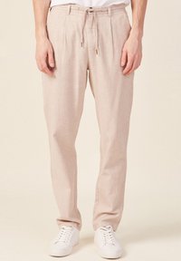 Pantalons en lin beige clair à coupe décontractée, dotés d'une taille élastique avec cordon de serrage, de plis et d'une texture lisse, associés à des baskets blanches.