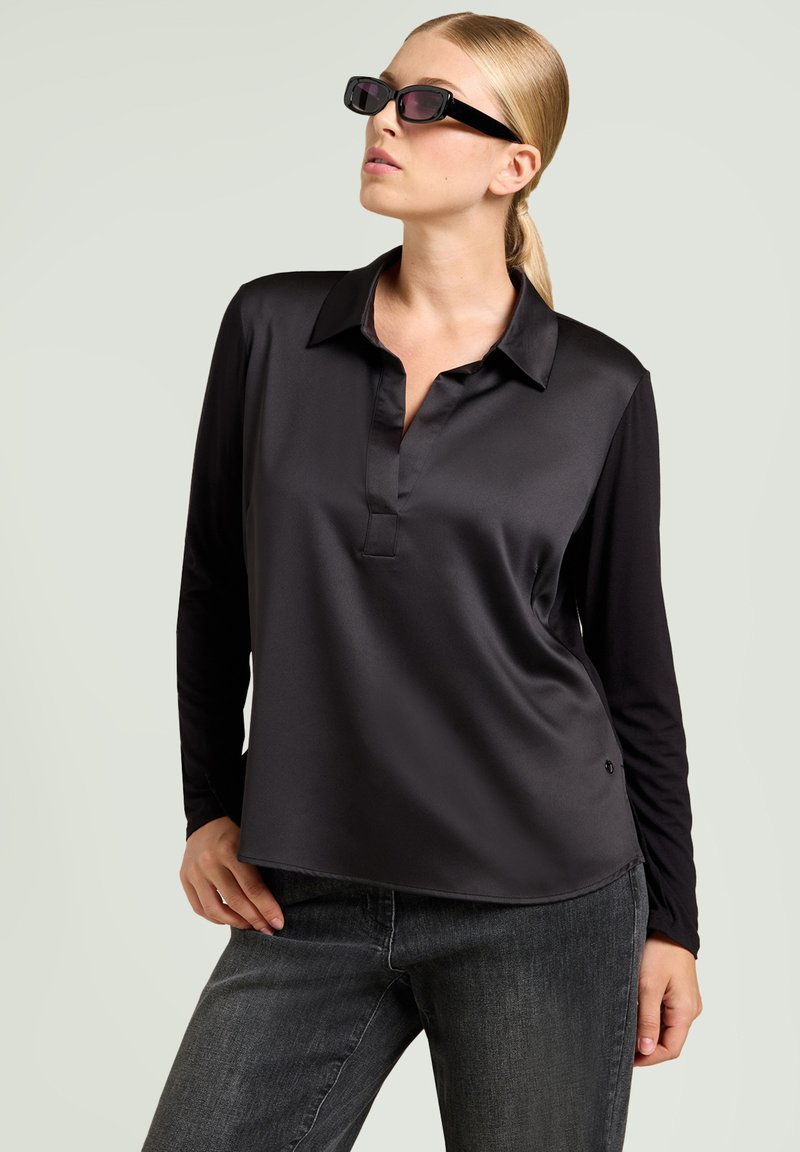 Polo shirt lungo in satin nero con colletto. Tessuto liscio e design semplice. Abbinato a pantaloni in denim grigi e occhiali da sole scuri.