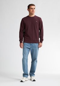 Bordeaux sweatshirt met lange mouwen, ronde hals en een eenvoudig logo. Gecombineerd met lichtblauwe jeans en witte sneakers.