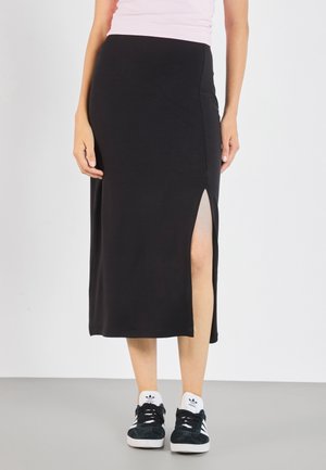 Vrouw draagt een zwarte midi-rok met een hoge split aan de dij en zwarte Adidas sneakers met witte zolen en strepen.