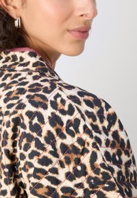 Steppjacke im Leopardenmuster mit Schwarz-, Beige- und Brauntönen. Verfügt über einen Kragen und kontrastierende Nähte. Weicher Stoff.