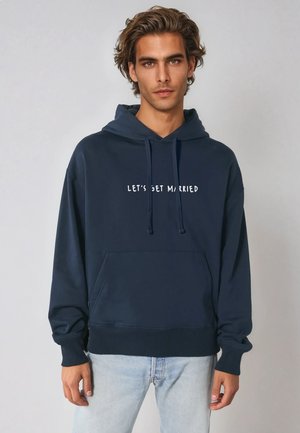 Marineblauwe hoodie van zacht materiaal, met een voorzak en de tekst "Laten we trouwen" in het wit. Comfortabele pasvorm met een capuchon met trekkoord.