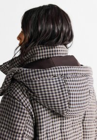 Gepolsterte Steppjacke in Brauntönen mit kariertem Muster. Verfügt über eine große Kapuze und strukturierte Stoffe, mit kontrastierenden einfarbigen Akzenten an den Schultern.