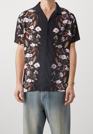 Mężczyzna ubrany w czarną koszulę na guziki z krótkim rękawem, ozdobioną nadrukiem tygrysa i różowych kwiatów, w połączeniu z wyblakłymi niebieskimi dżinsami i kolorową bransoletką z koralików.