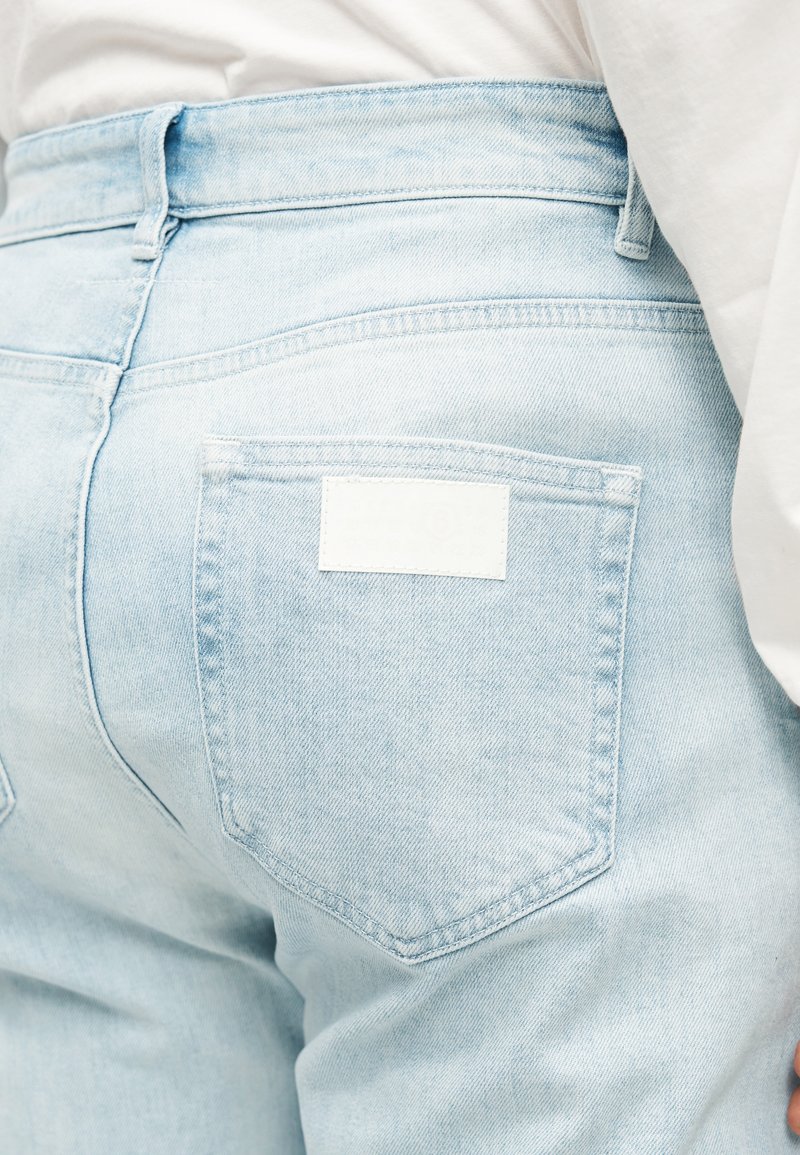 Lyseblå denimjeans med en baglomme og et tomt hvidt mærke. Markante blå sømme langs syningerne, glat tekstur.