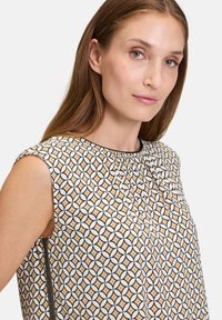 Mönstrat ärmlöst linne med en geometrisk design i beige, vitt och svart, med en veckad halsringning och sidoglittrad detalj.