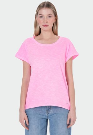 Jonge vrouw met steil blond haar die een felroze korte mouwen T-shirt en lichtblauwe spijkerbroek draagt, staat voor een effen achtergrond.