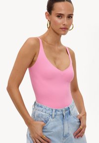 Femme portant un haut sans manches rose et un jean taille haute bleu clair, avec des boucles d'oreilles anneaux dorés et des bagues, posant les mains dans les poches.