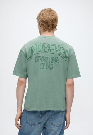 Blond person set bagfra iført en grøn t-shirt med teksten "LINDBERGH SPORTING CLUB" på ryggen og blå cowboybukser.