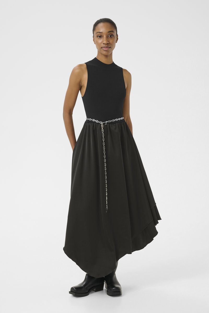 Haut sans manches noir à côtes associé à une jupe noire fluide, avec une ceinture en chaîne argentée et des bottes à talons noires. Tissu lisse avec un ourlet en couches.