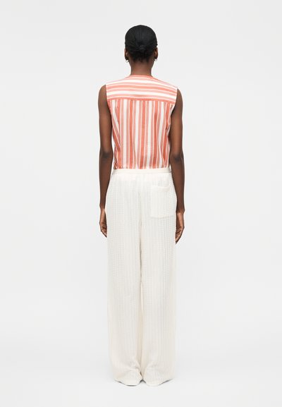 Γυναίκα με μαλλιά σε χαμηλό κότσο, που φοράει αμάνικο πορτοκαλί-λευκό μπλουζάκι με κάθετες ρίγες και φαρδιά άσπρη υφασμάτινη παντελόνα, στέκεται με την πλάτη στο φακό.