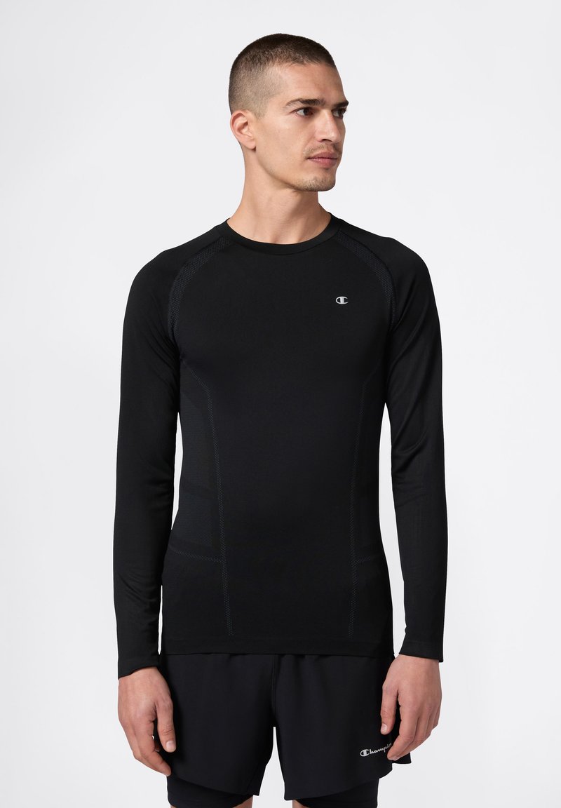 Mann in einem enganliegenden schwarzen Langarm-Sportshirt und schwarzen Shorts, der steht und nach rechts schaut, vor einem weißen Hintergrund.