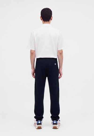 Homme debout de dos, portant une chemise blanche à manches courtes, un pantalon noir avec un petit logo et des baskets blanches avec un motif de feuille d’érable bleue.