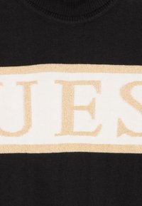 Črna pulover s belim horizontalnim trakom in zlatim napisom "GUESS". Mehka pletenina z okroglim izrezom in rebrastimi robovi.