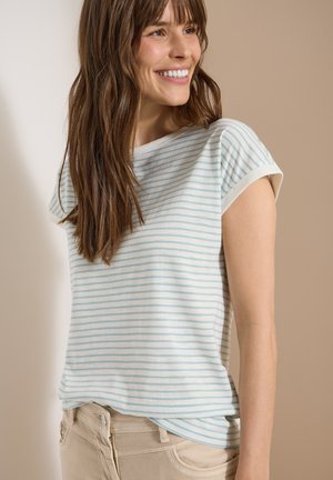 Young woman with long brown hair smiling, wearing a white and light blue striped short-sleeve shirt and beige pants, standing against a plain background.