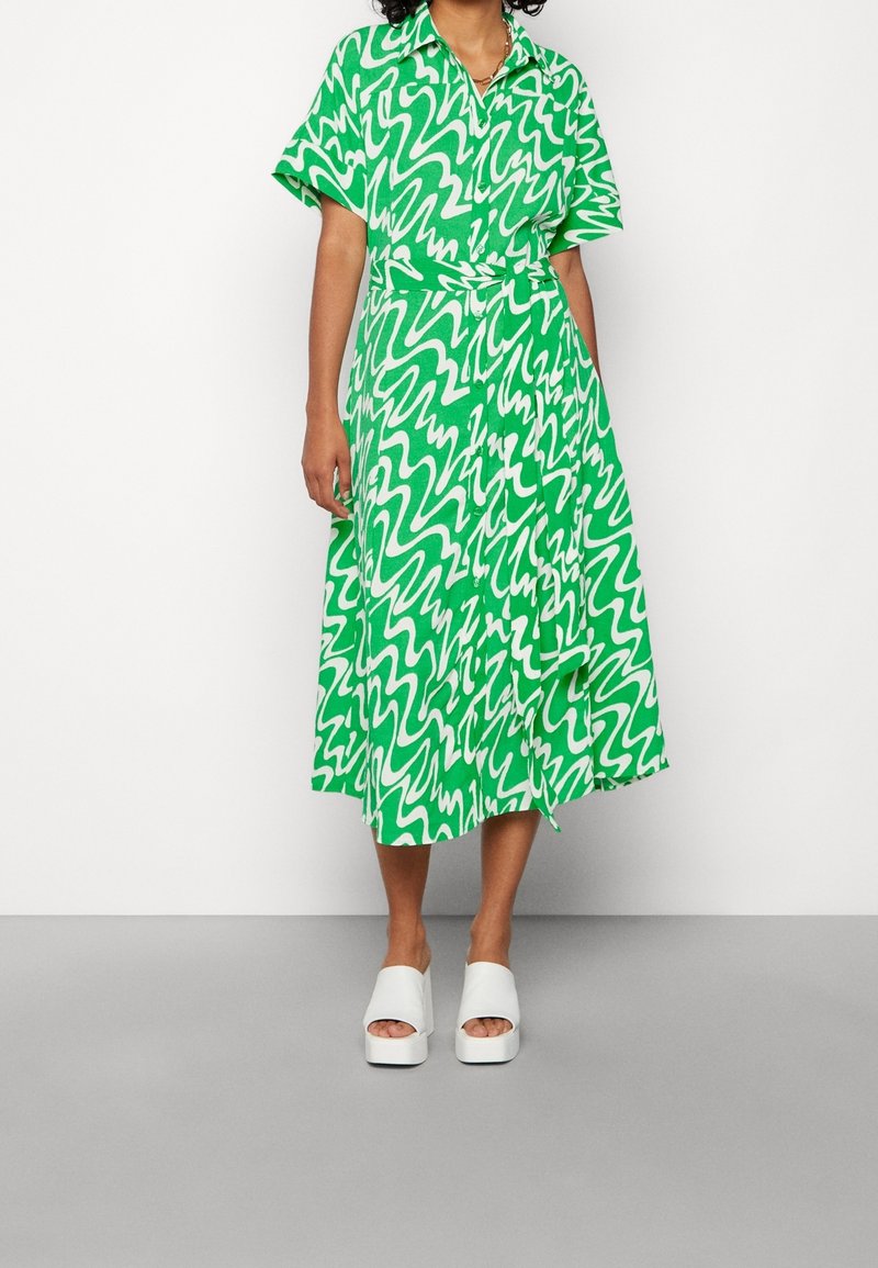 Personne portant une robe midi à motif abstrait vert et blanc, avec manches courtes et ceinture, et des sandales compensées blanches à bout ouvert.