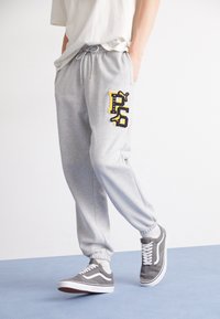 Grå sweatpants med elastisk talje og snøre, samt et sort og gult P-logo; parret med grå sneakers.