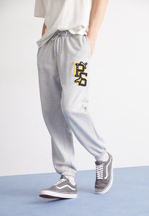 Grå sweatpants med elastisk talje og snøre, samt et sort og gult P-logo; parret med grå sneakers.