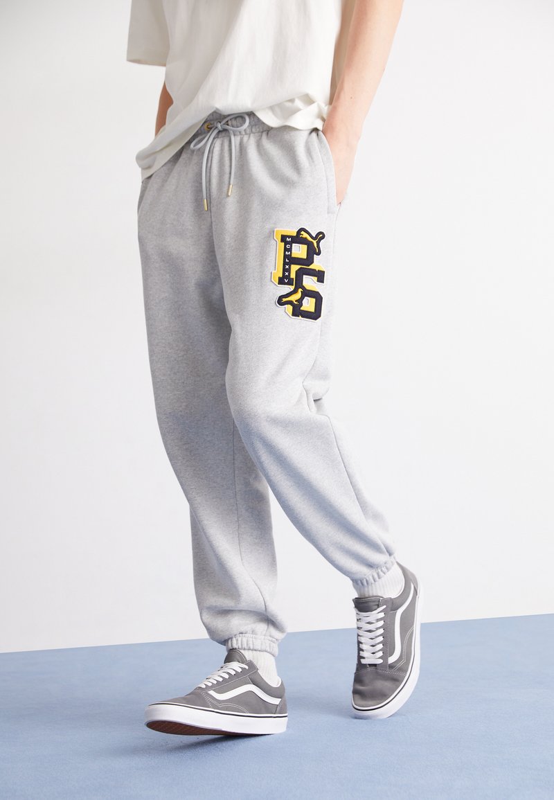 Grå sweatpants med elastisk talje og snøre, samt et sort og gult P-logo; parret med grå sneakers.