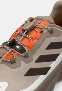 Lys brun joggesko med oransje aksent, teksturert stoff, sikkert snøresystem og tre mørke striper på siden.