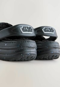 Zwarte instapsandalen met een fuzzy textuur, met een ronde "Star Wars" logo op de banden. Getextureerde zool met kleurrijke spikkels.