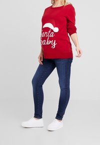 Red sweater with "Santa Baby" text and white Santa hat design, paired with dark blue skinny jeans and white sneakers. Soft fabric texture.