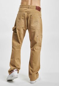 Brune cargo-bukser med flere lommer, syningdetaljer og et logo-patch på den bageste talje. Lavet af bomuldsstof, løs pasform og bred ben-design.