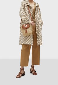 Person iført beige og brunternet jakke, beige bukser med vide ben, brune sandaler med hæl, og bærer en flettet skuldertaske med kvast og bogstavet M.
