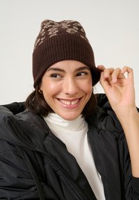 Bruine gebreide beanie met een ribbelstructuur en een bloemenpatroon in lichtbruin. Draagt het met een zwarte gewatteerde jas en een witte coltrui eronder.