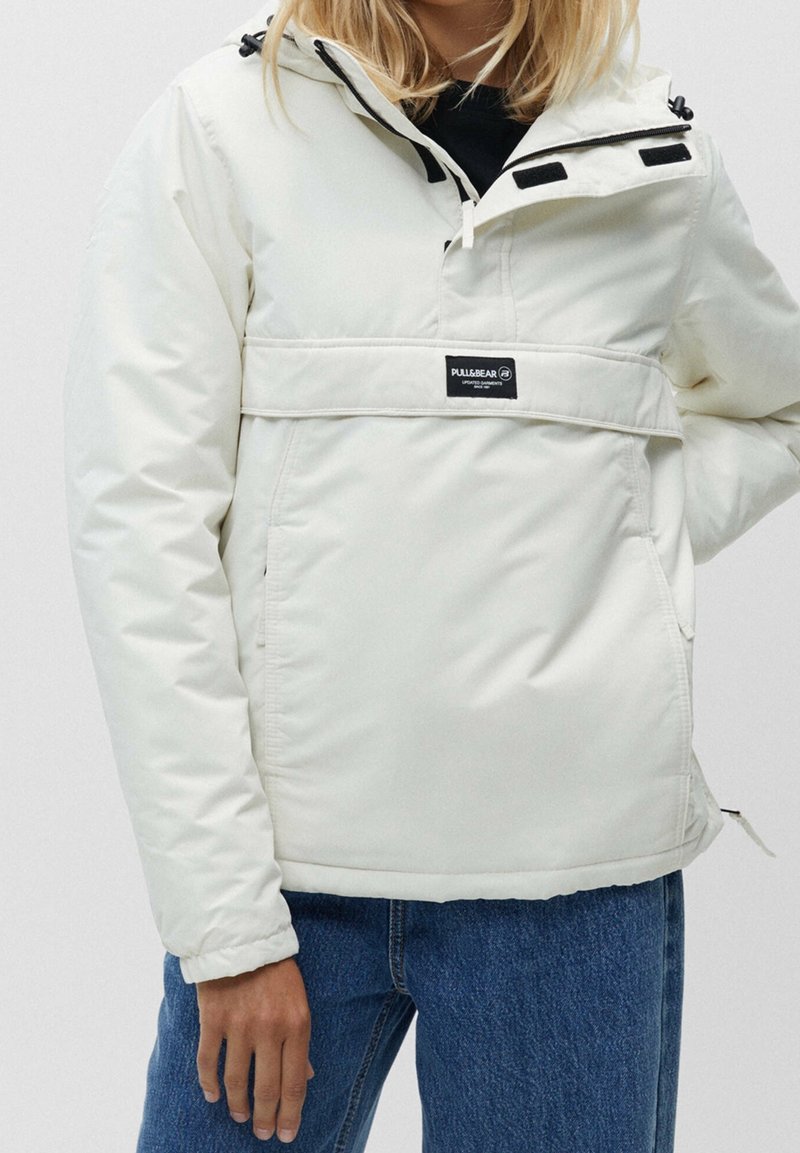 Lättviktig vit pullover-jacka med huva, med en framficka, svarta detaljer och elastiska mudd. Materialet verkar slätt och syntetiskt.