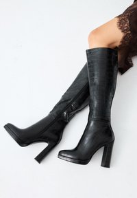 Botas negras hasta la rodilla hechas de cuero brillante con estampado de cocodrilo. Punta cuadrada, tacón alto y bloque, y cremallera trasera para el cierre.