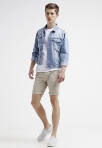 Hellblaue Jeansjacke mit zwei Brusttaschen, weißem Rundhalsausschnitt-T-Shirt, beigen Shorts und weißen Slippern mit durchbrochenem Muster.
